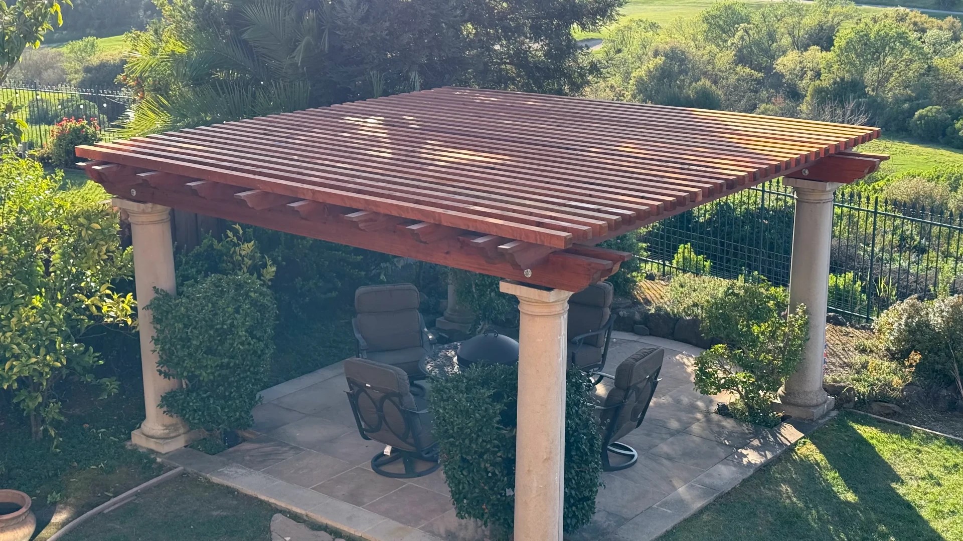 New redwood pergola top with shaped rafter tails and slats on four stone columns over a patio with chairs