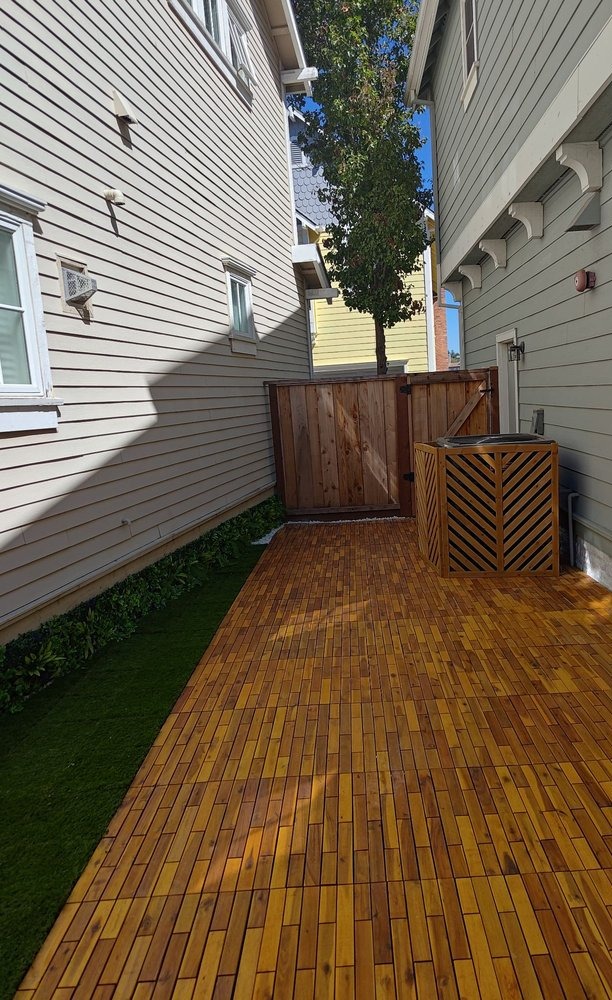 Narrow side yard finished with wood deck tiles, a strip of artificial turf, a slatted AC screen and a wood gate