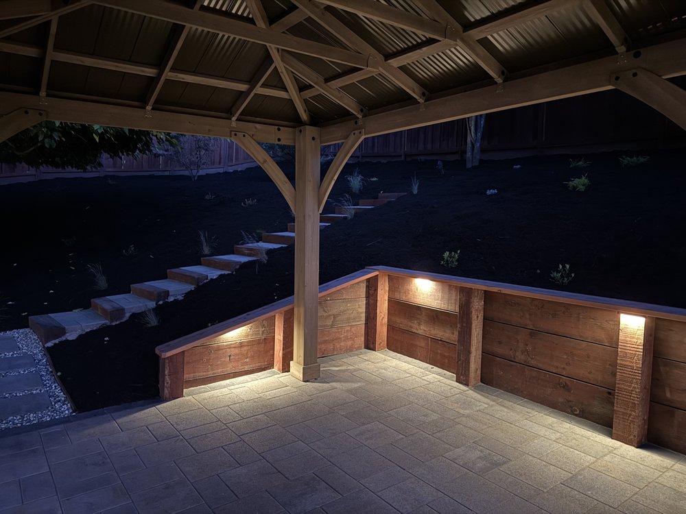 View from under the lit gazebo across the paver patio toward the wood retaining wall and the hillside steps