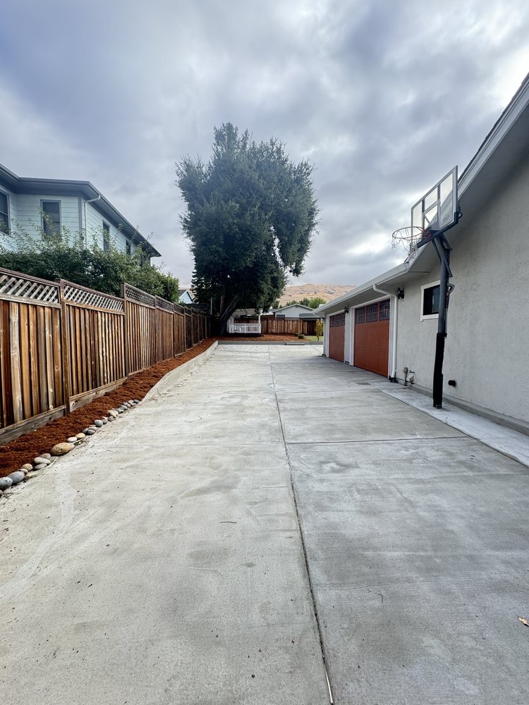 Finished wide concrete driveway running alongside the garage in Milpitas, with a new redwood fence and cobble-edged bark border on the far side