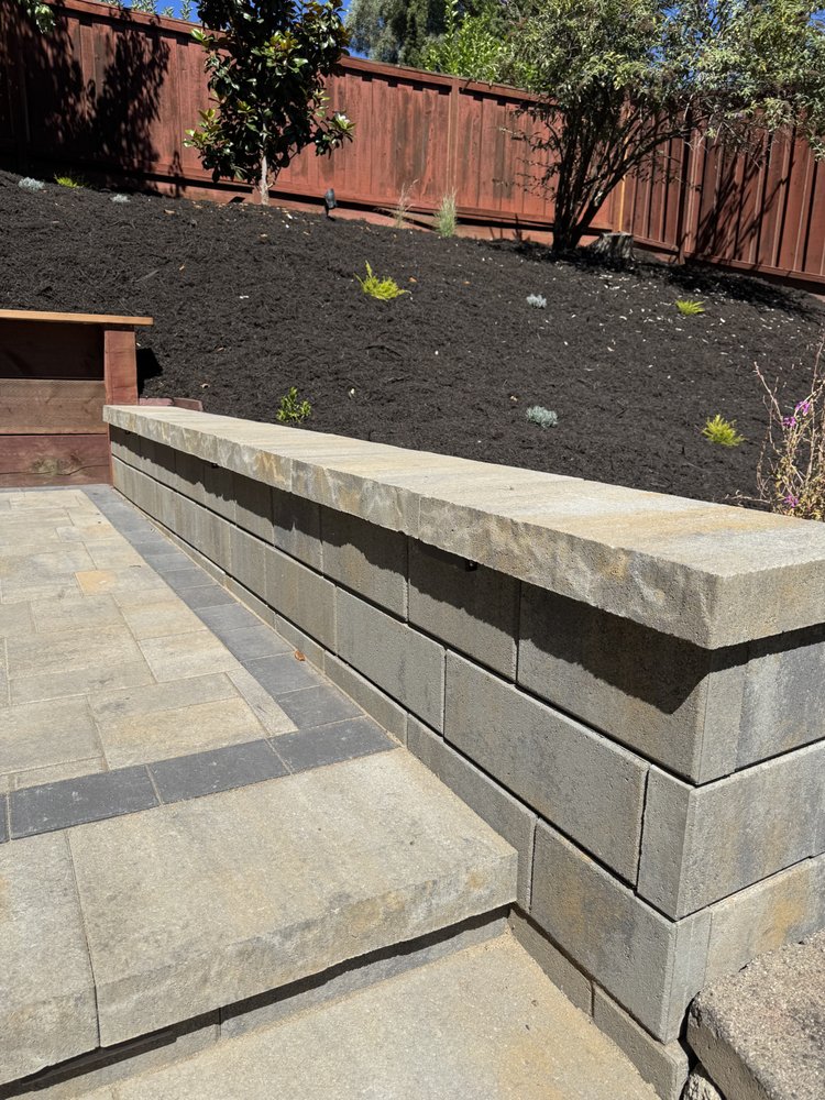 Block retaining wall with a heavy cut-stone cap running alongside the paver patio, holding back the planted hillside